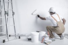 a man paints a white wall with a roller. Repair of the interior. Young male decorator painting a wall in the empty room, concept builder or painter in helmet with paint roller over the empty room.